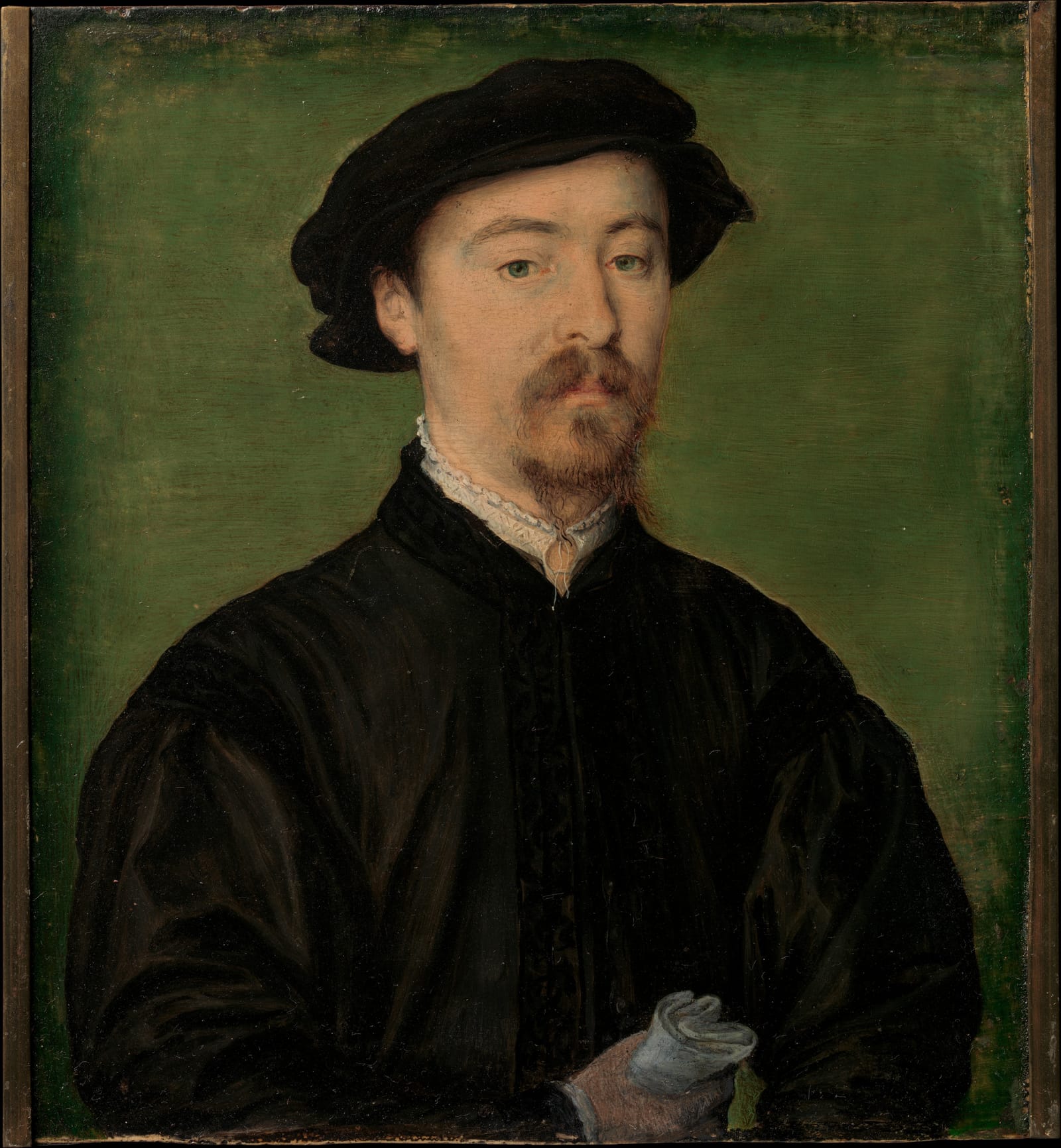 A bearded man in a black cap and coat holds a pair of gloves against a green ground.